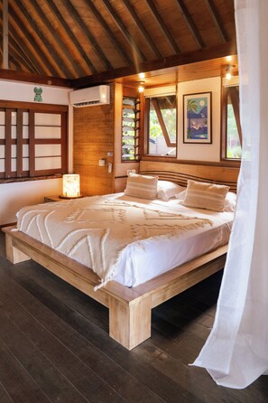A wooden-framed bed with a white bedspread and pillows, a wall-mounted air conditioner, and a window with a view of greenery.
