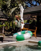 A person standing by a pool with a green inflatable ring, a wooden deck, and a covered outdoor seating area.
