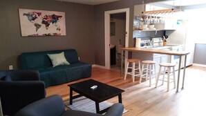 A living room with a sofa, a coffee table, and a kitchen area with stools. - Wine Alley East suite (Hermann)