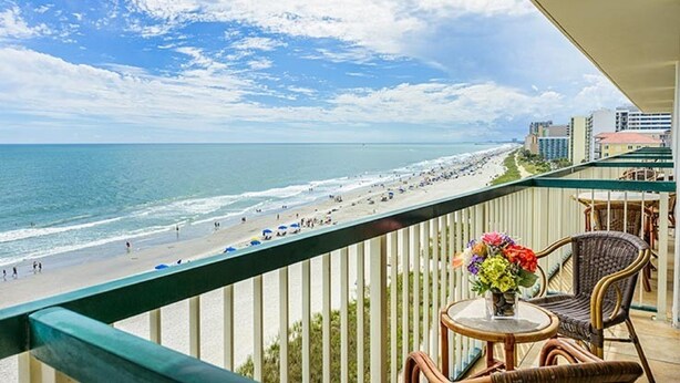 A balcony with a view of a beach and ocean, wicker furniture, and a vase with flowers. -  Special Exceptional Offer Guaranteed Oceanfront Villa  Available July 11-18 (Myrtle Beach)