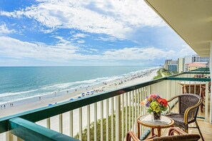 A balcony with a view of a beach and ocean, wicker furniture, and a vase with flowers.