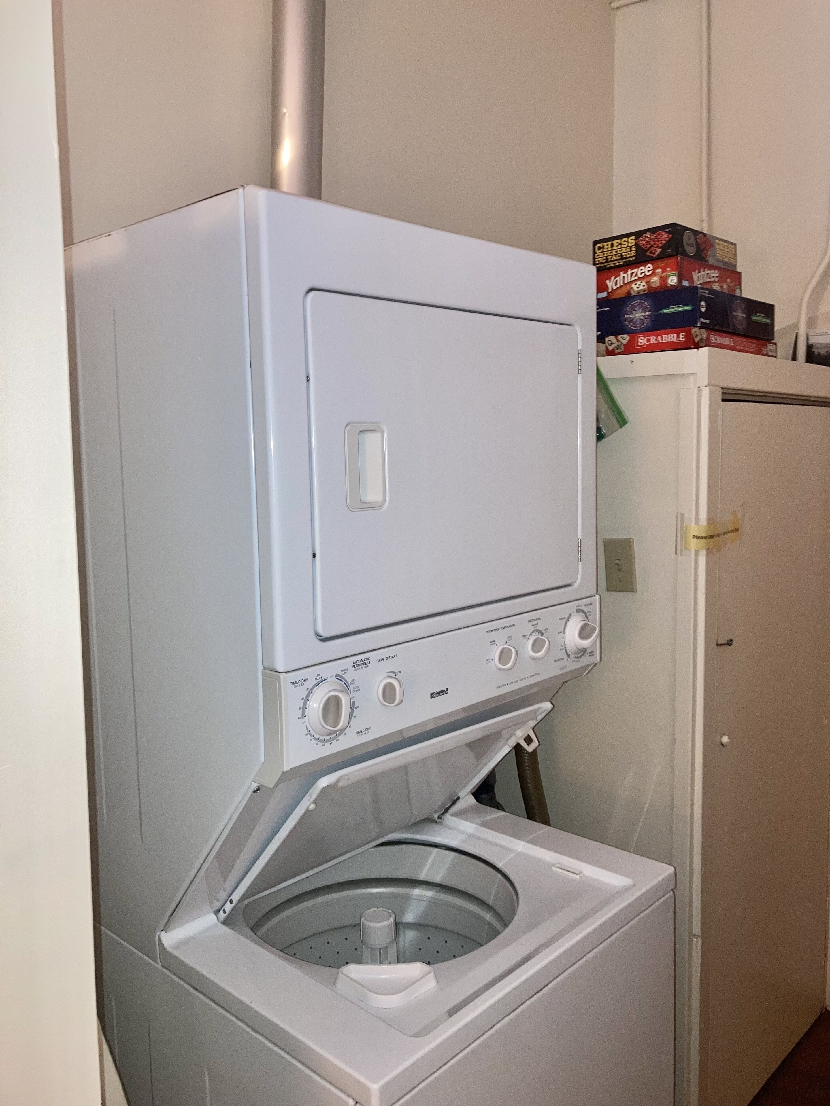A compact, white front-loading washing machine with a front-loading dryer, located in a small laundry room with a white door and a shelf above holding board games.