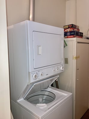 Una rentadora blanca i compacta de càrrega frontal, amb un assecadora de càrrega frontal, situada en un petit espai de bugaderia amb una porta blanca i una estant a la part superior on hi ha jocs de taula.