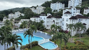 Ein Resort mit Pool, umgeben von Palmen und roten Blüten, vor einer Kulisse weißer Gebäude mit Balkonen.