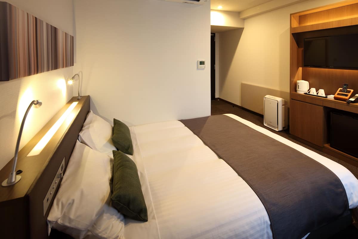 Habitación de hotel con cama, mesita de noche, lámpara y una pequeña zona de kitchenette equipada con microondas y cafetera.