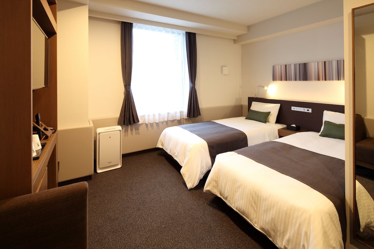 Habitación de hotel con dos camas, un escritorio, una silla, un ventanal con cortinas y una lámpara de pared.