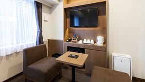 A compact hotel room with a television mounted on a wooden wall, a small table with a phone and a notebook, two armchairs, a coffee maker, and a mini-fridge.