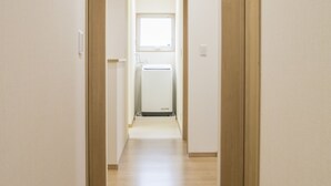A narrow hallway with wooden flooring and light-colored walls, leading to a room with a window and a white appliance. - Koko (Fujikawaguchiko)
