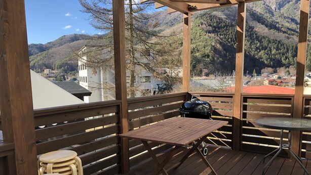 A wooden deck with a table and chairs, a grill, and a view of a mountainous landscape. - Koko (Fujikawaguchiko)