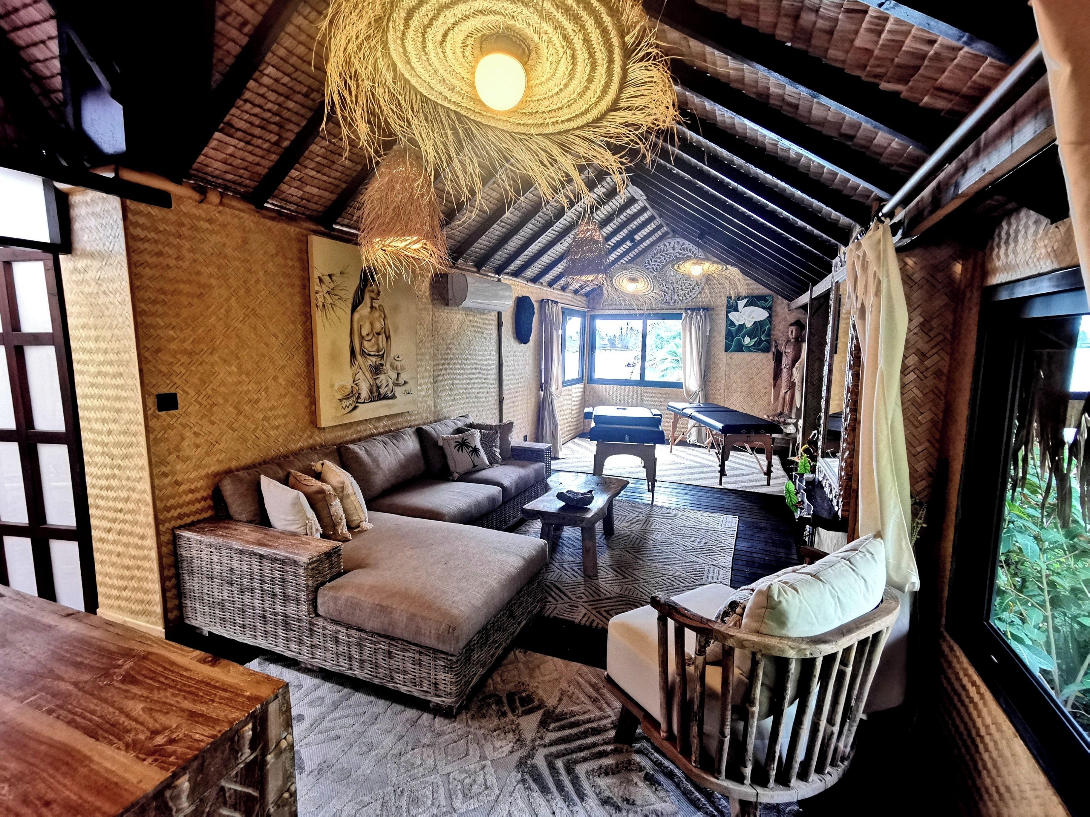 A cozy room with a sofa, a wicker chair, a massage table, and a large window with a view of greenery.