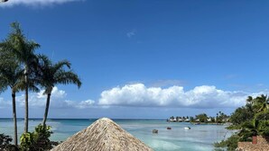Escena de playa tropical con palmeras, agua azul cristalina y una cabaña con techo de paja.