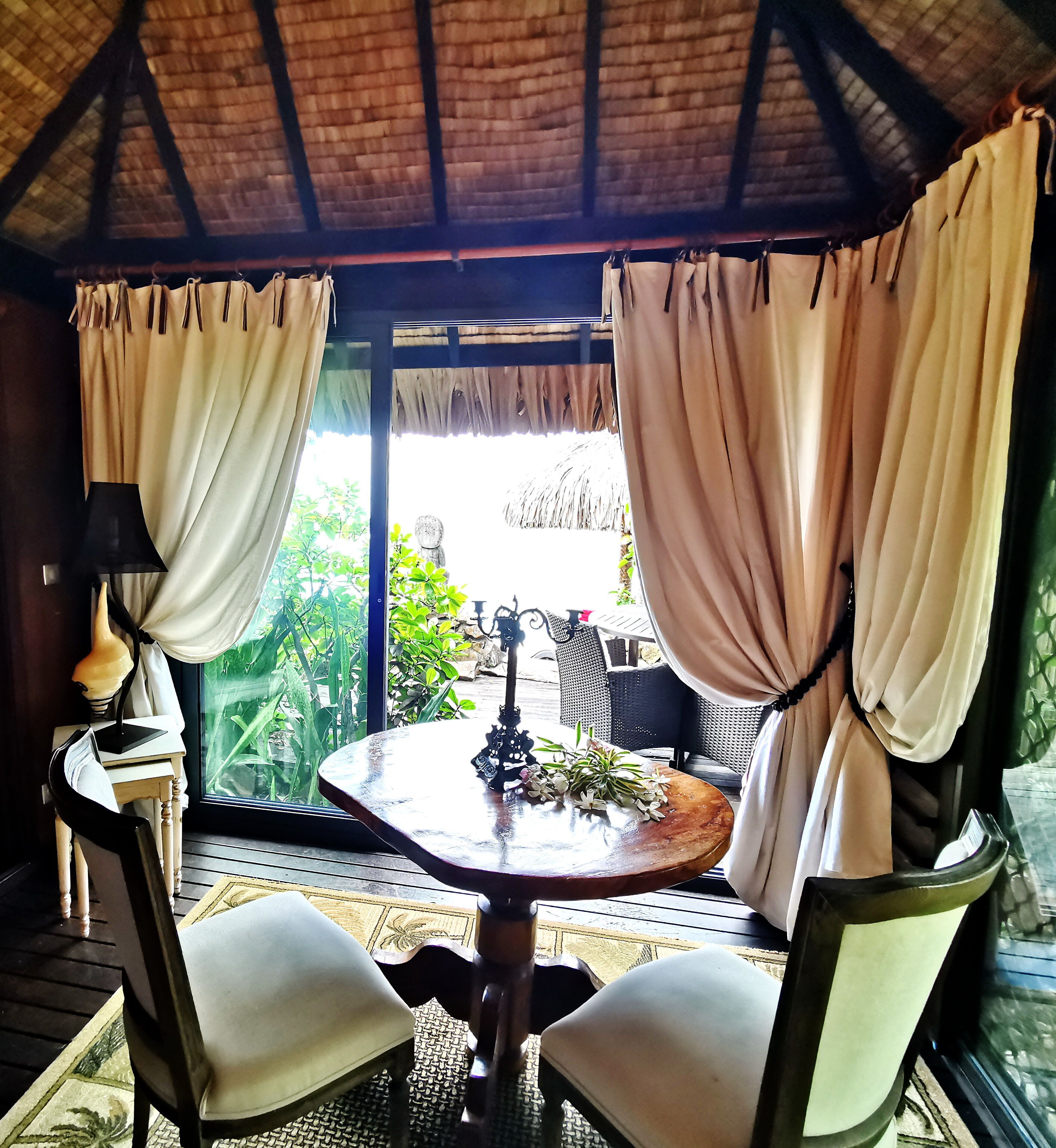 A covered outdoor seating area with a table and chairs, a decorative lamp, and a view of greenery outside.