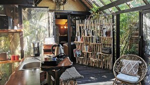 Una cocina rústica y acogedora con mostrador de madera, una máquina de café y un hervidor. Hay una silla de mimbre con cojín y una estantería llena de libros y objetos decorativos.