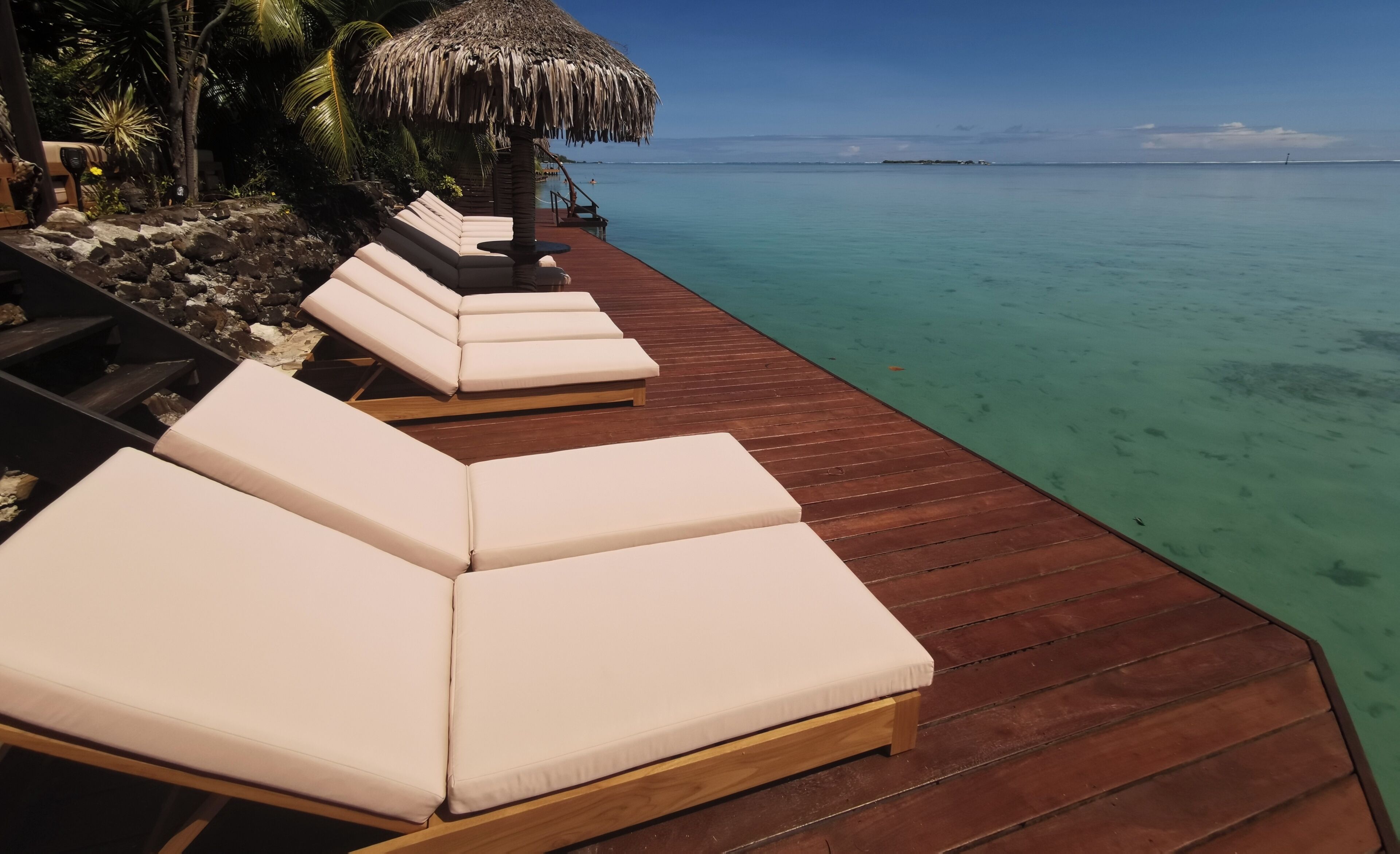 A wooden deck with white cushioned lounge chairs overlooking turquoise waters.
