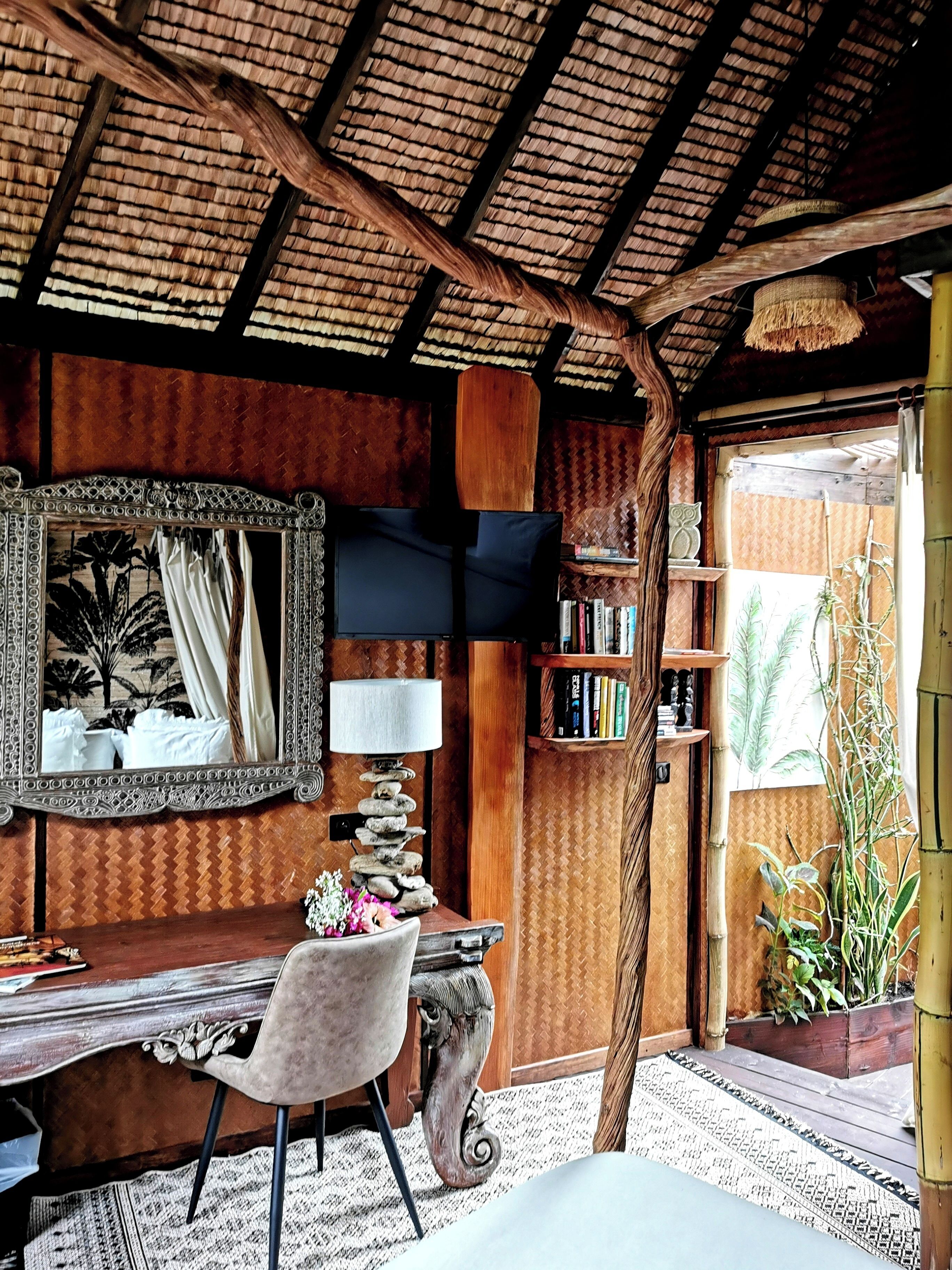 A cozy room with a wooden ceiling, a desk with a chair, a mirror, a lamp, and a bookshelf.
