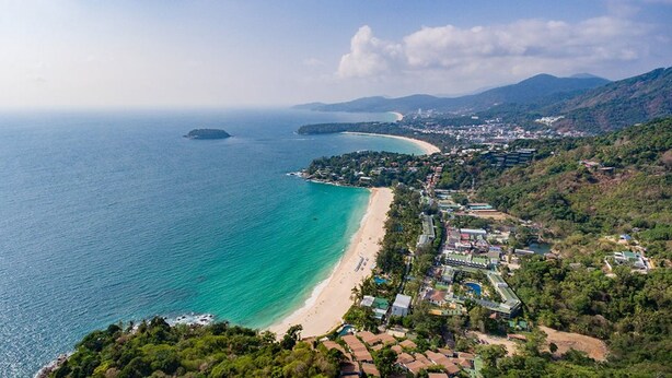 An aerial view of a coastal resort with a long sandy beach, a small island, and a mountainous backdrop.