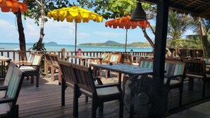An outdoor dining area with wooden tables and chairs, umbrellas, and a view of the sea.