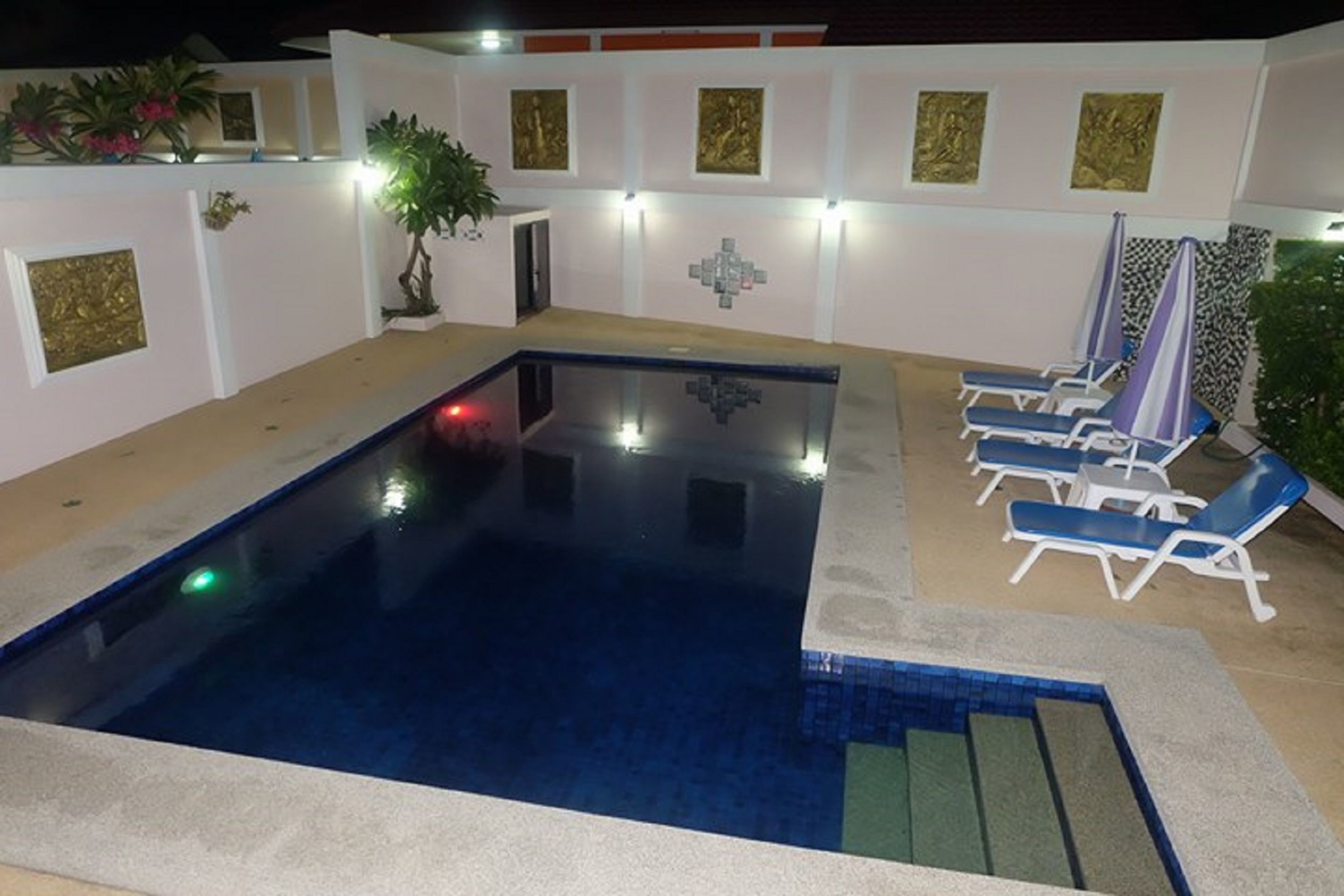 A swimming pool with a blue interior, surrounded by white walls with gold accents and a tiled border. There are lounge chairs and a potted plant near the pool.