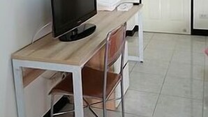 A room with a wooden desk, a television, a chair, a white door, and a tiled floor.