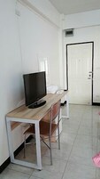 Una habitación con un escritorio de madera, televisor, una silla, una puerta blanca y piso de baldosas.