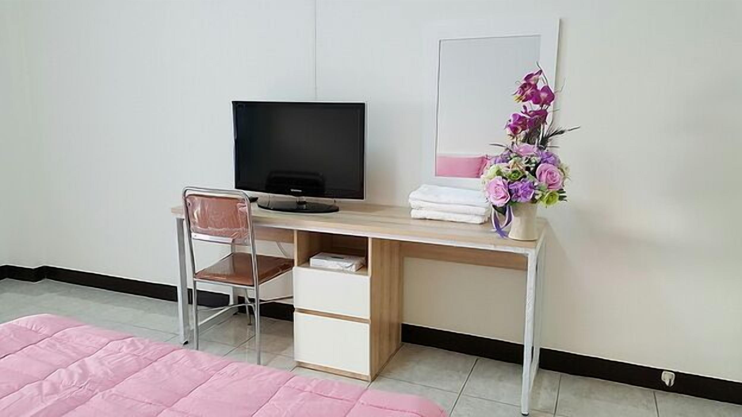 Un dormitorio con un escritorio de madera, una silla, un televisor, un espejo, una cama rosa y un florero con flores.