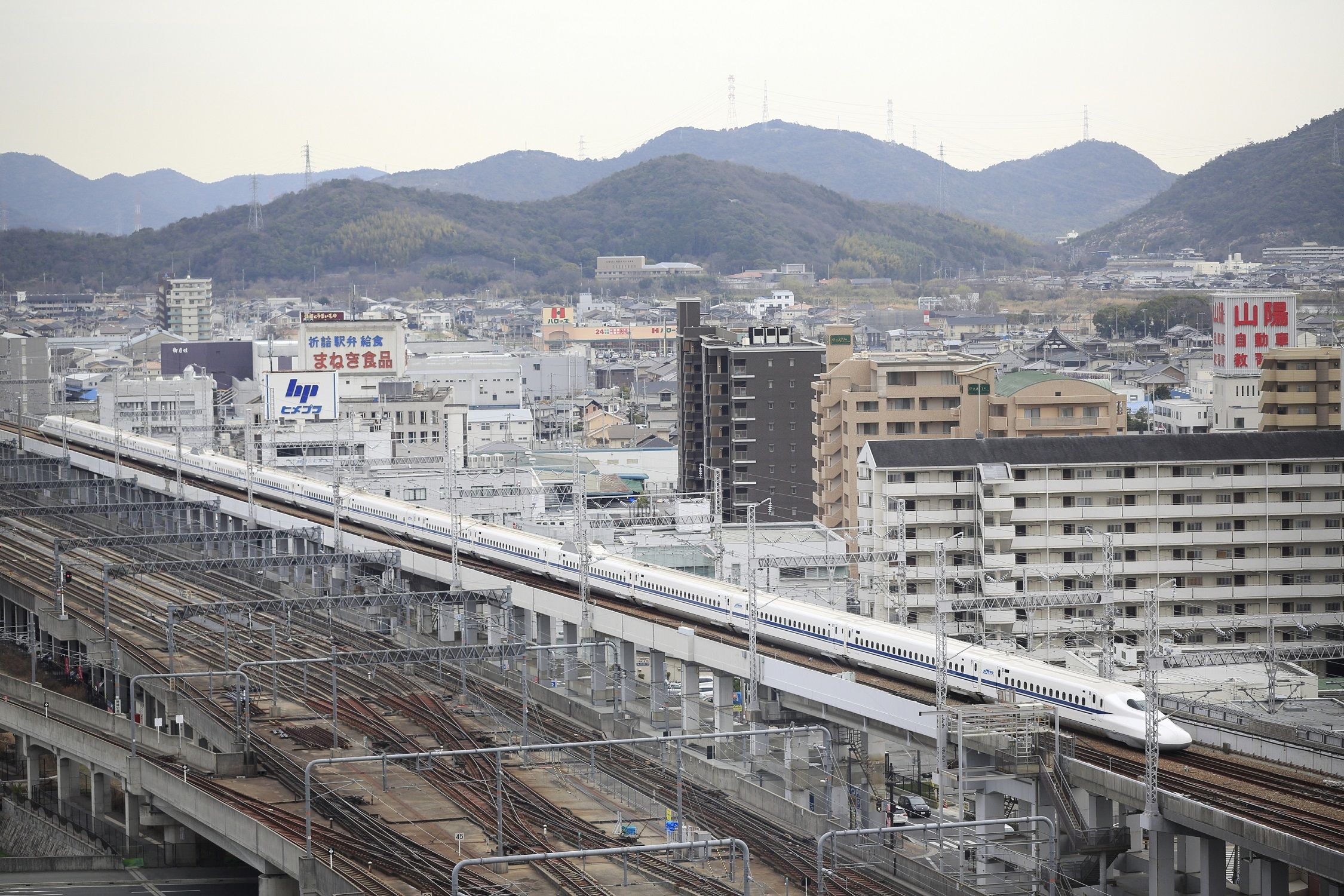 A train station with multiple tracks and a train on the right. There are buildings and mountains in the background.