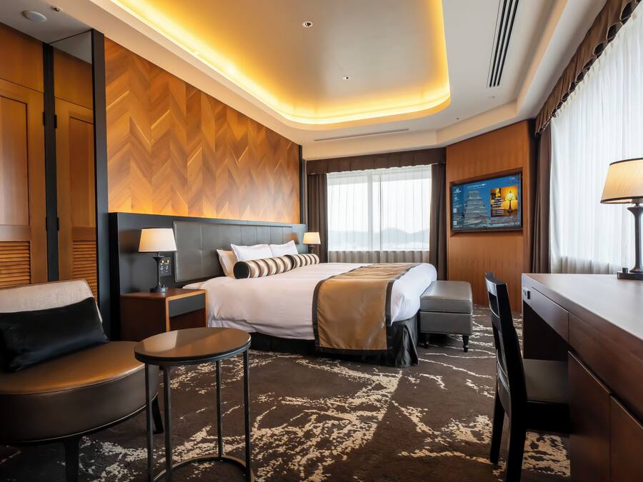 A hotel room with a large bed, a desk, a chair, a small table, and a TV on the wall.