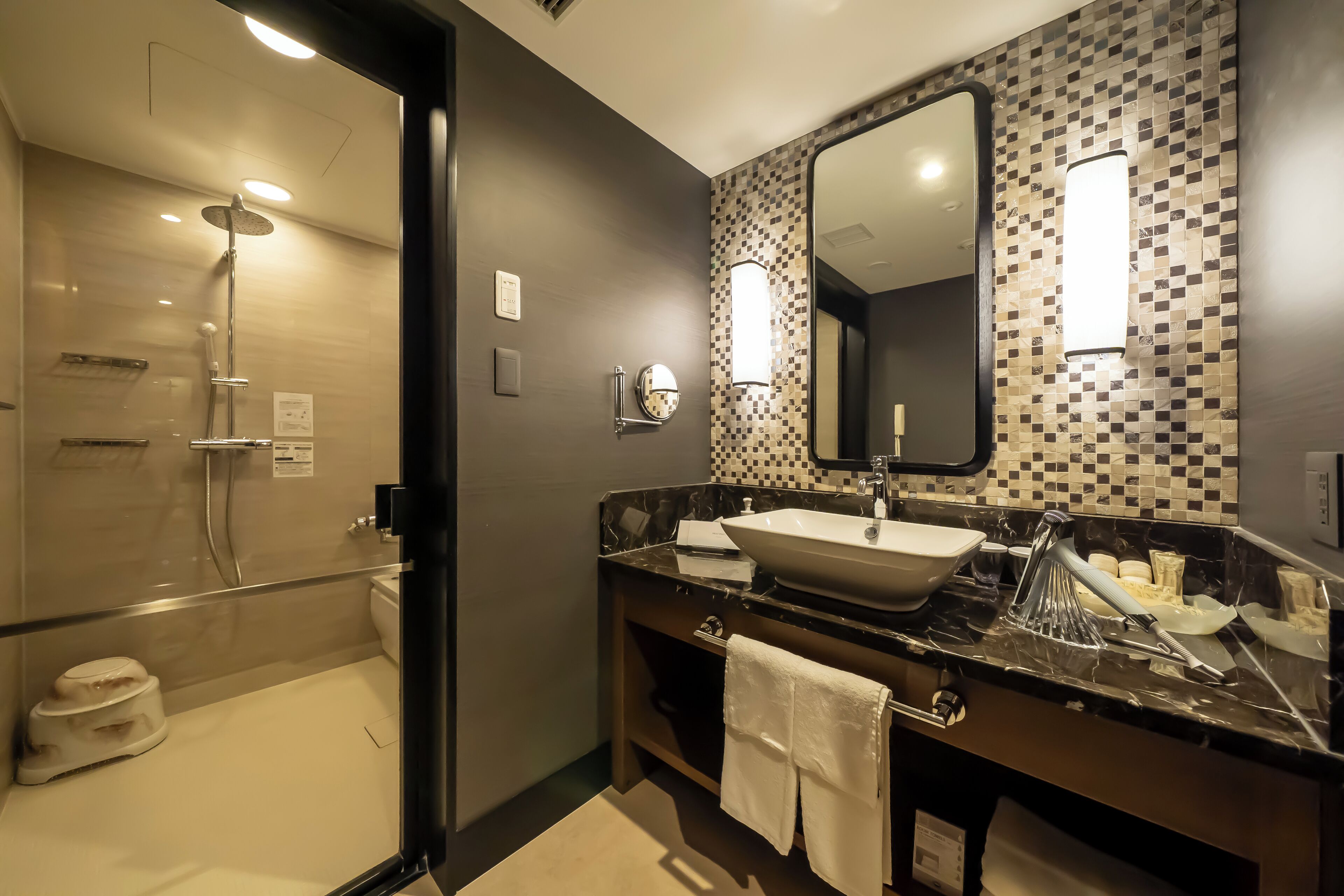A modern bathroom with a glass-enclosed shower, a large mirror, and a marble countertop with a sink and toiletries.