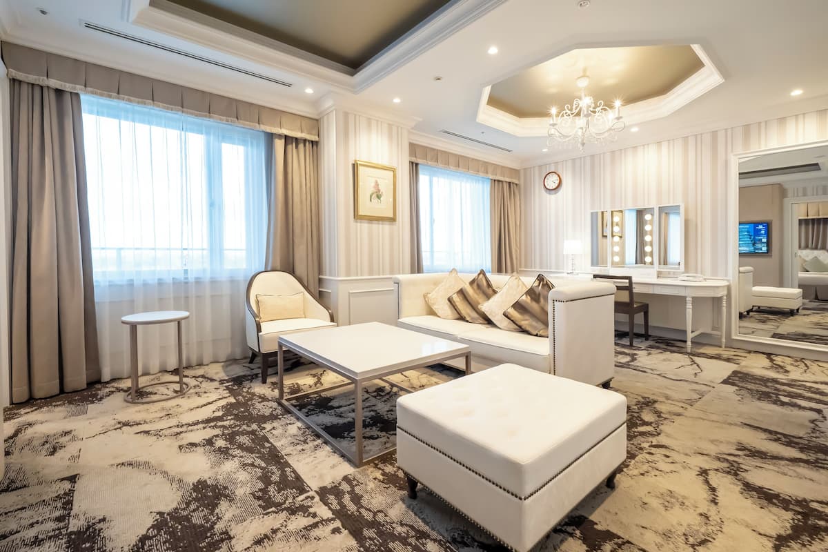 A spacious living room with a large sofa, a coffee table, and a small side table. There is a large window with curtains, a chandelier, and a mirror with lights.