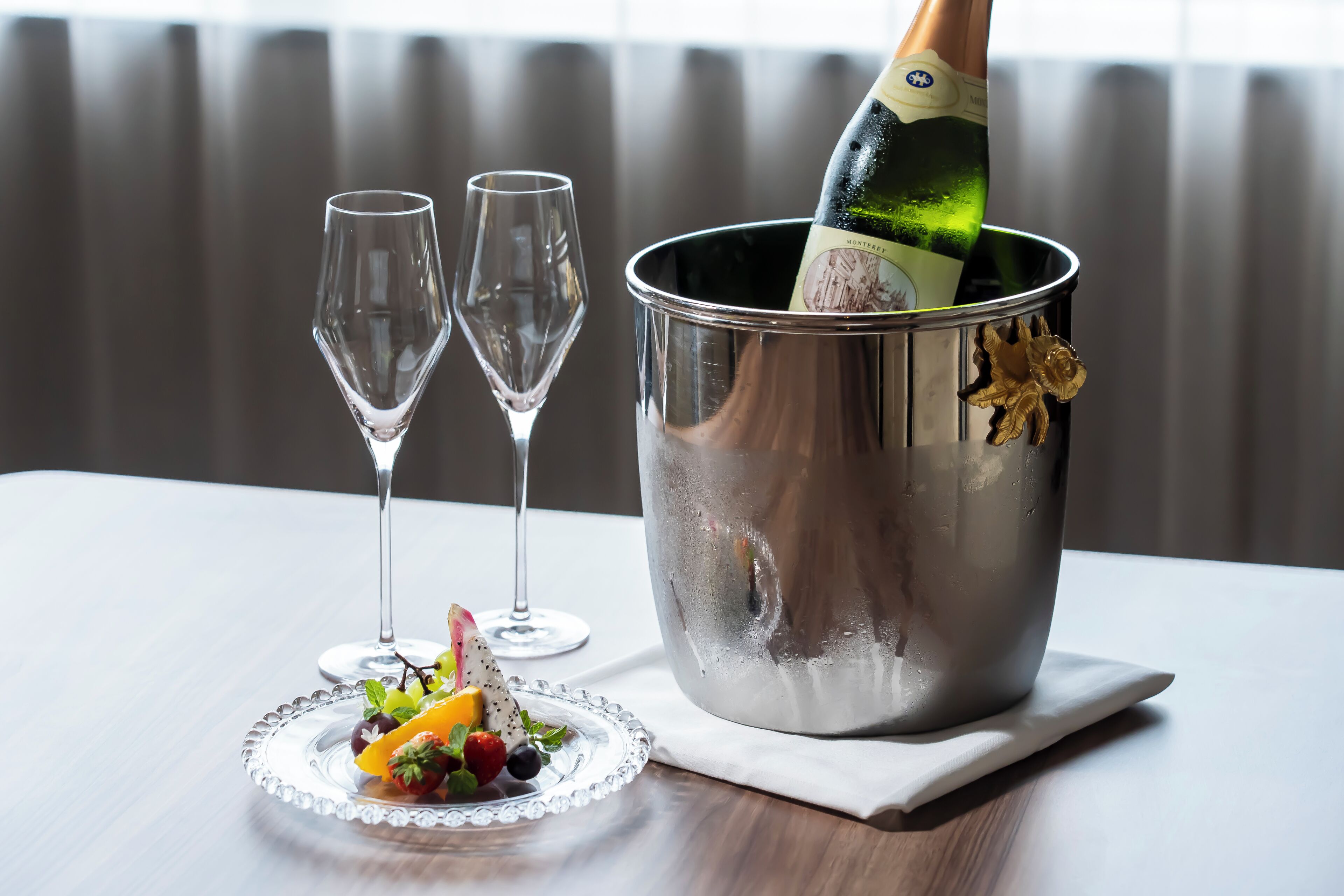 A champagne bottle in a metallic bucket with two empty glasses and a fruit platter on a table.