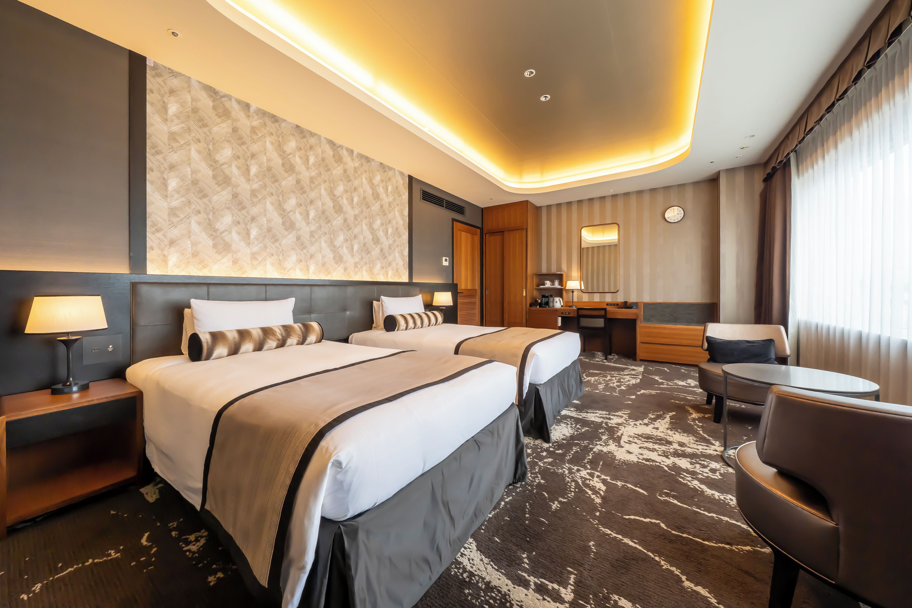 A hotel room with two beds, a desk, a chair, a lamp, and a large window.