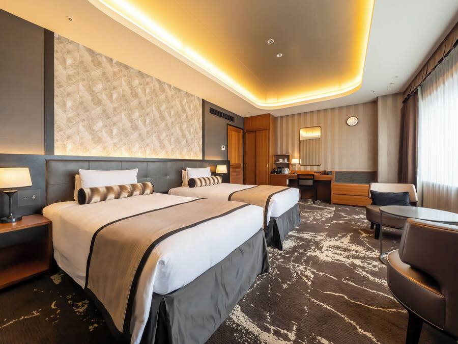 A hotel room with two beds, a desk, a chair, a lamp, and a large window.