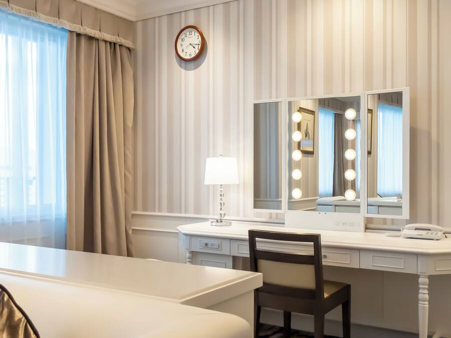 A hotel room with a bed, a desk with a mirror, a lamp, and a clock on the wall.