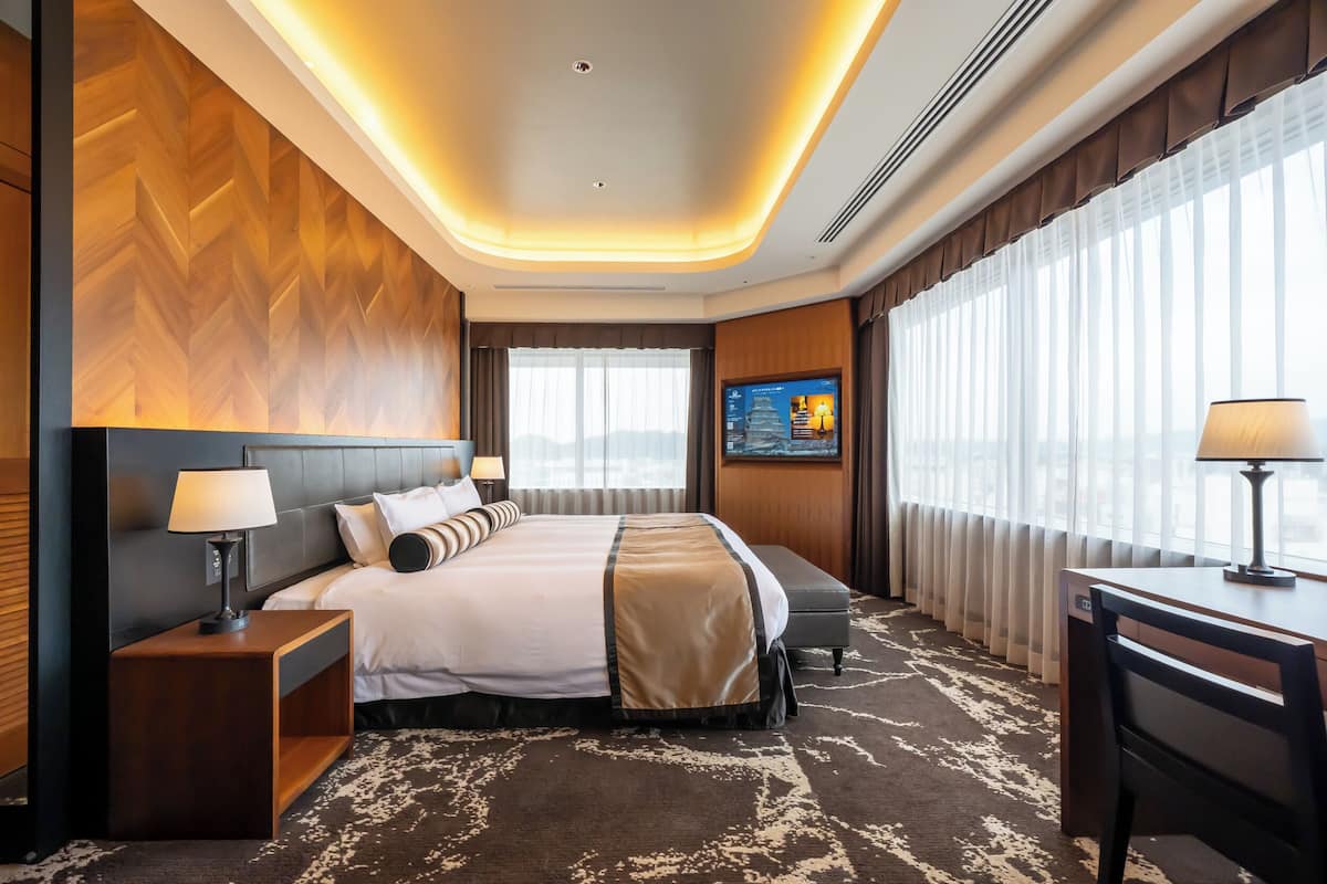 A hotel room with a large bed, bedside tables, a desk, and a view of the city through large windows.