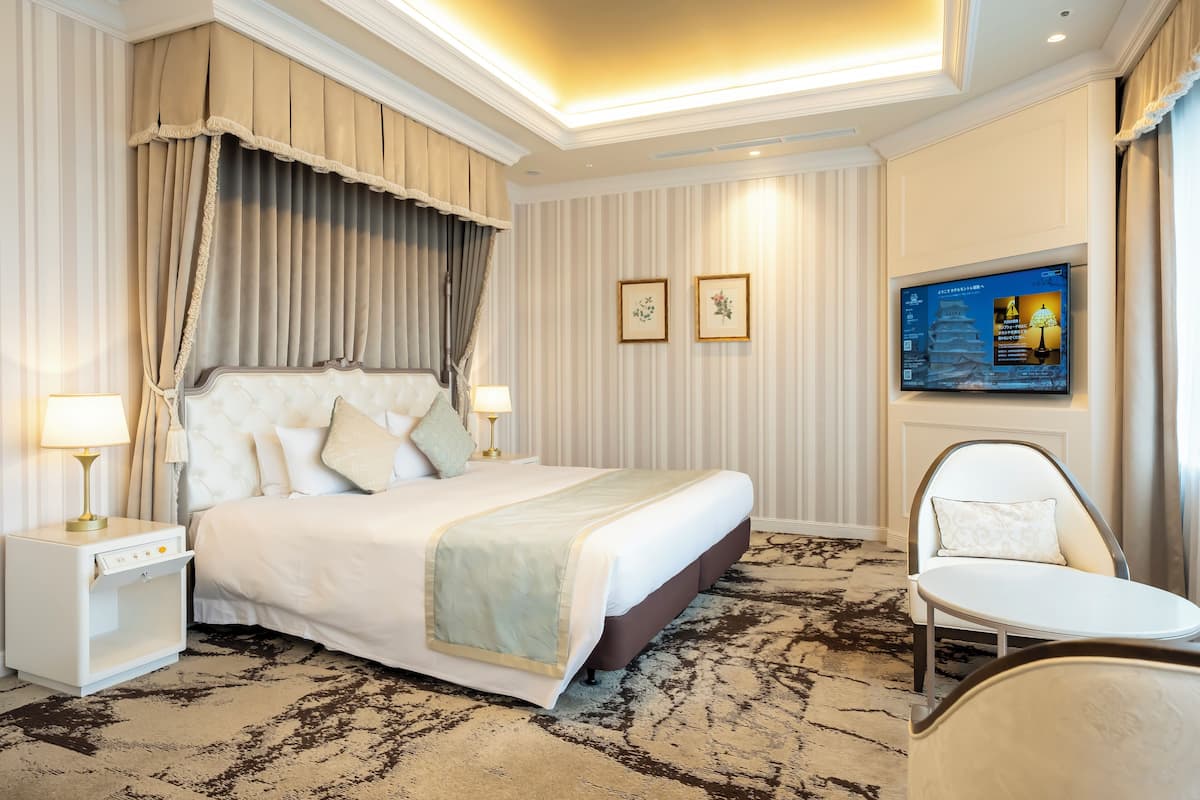 A hotel room with a large bed, a bedside table with a lamp, a chair, and a television mounted on the wall.
