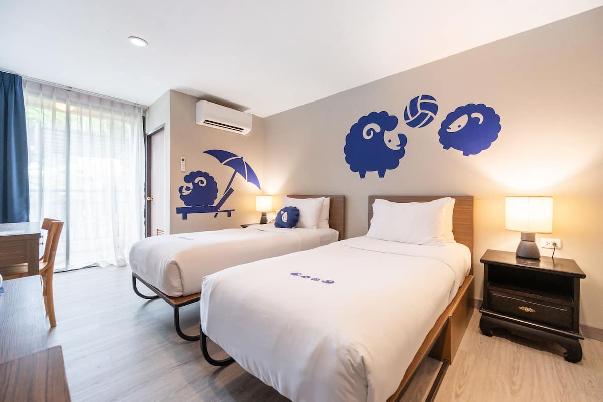 Habitación de hotel con dos camas, un escritorio y una silla. Decoración en la pared con motivos de ovejas y una escena de playa.