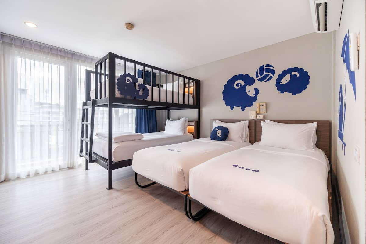 Una habitación de hotel con una cama nido, dos camas individuales, un ventanal con cortinas y adornos en la pared.