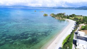 A coastal area with a beach, clear water, and a road leading to a white building. - Suite villa Yagaji (Nago)