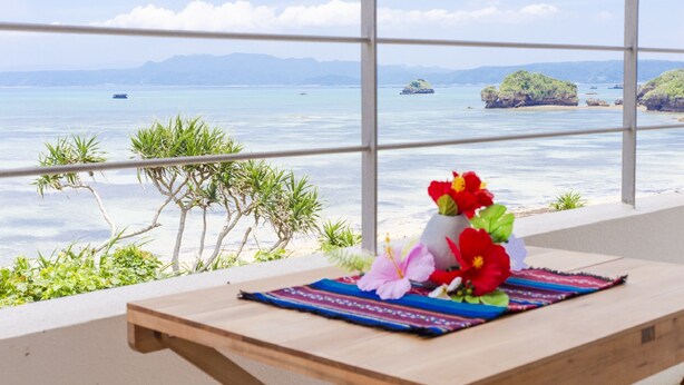 A balcony with a wooden table, a colorful woven mat, and a floral arrangement. - Suite villa Yagaji (Nago)