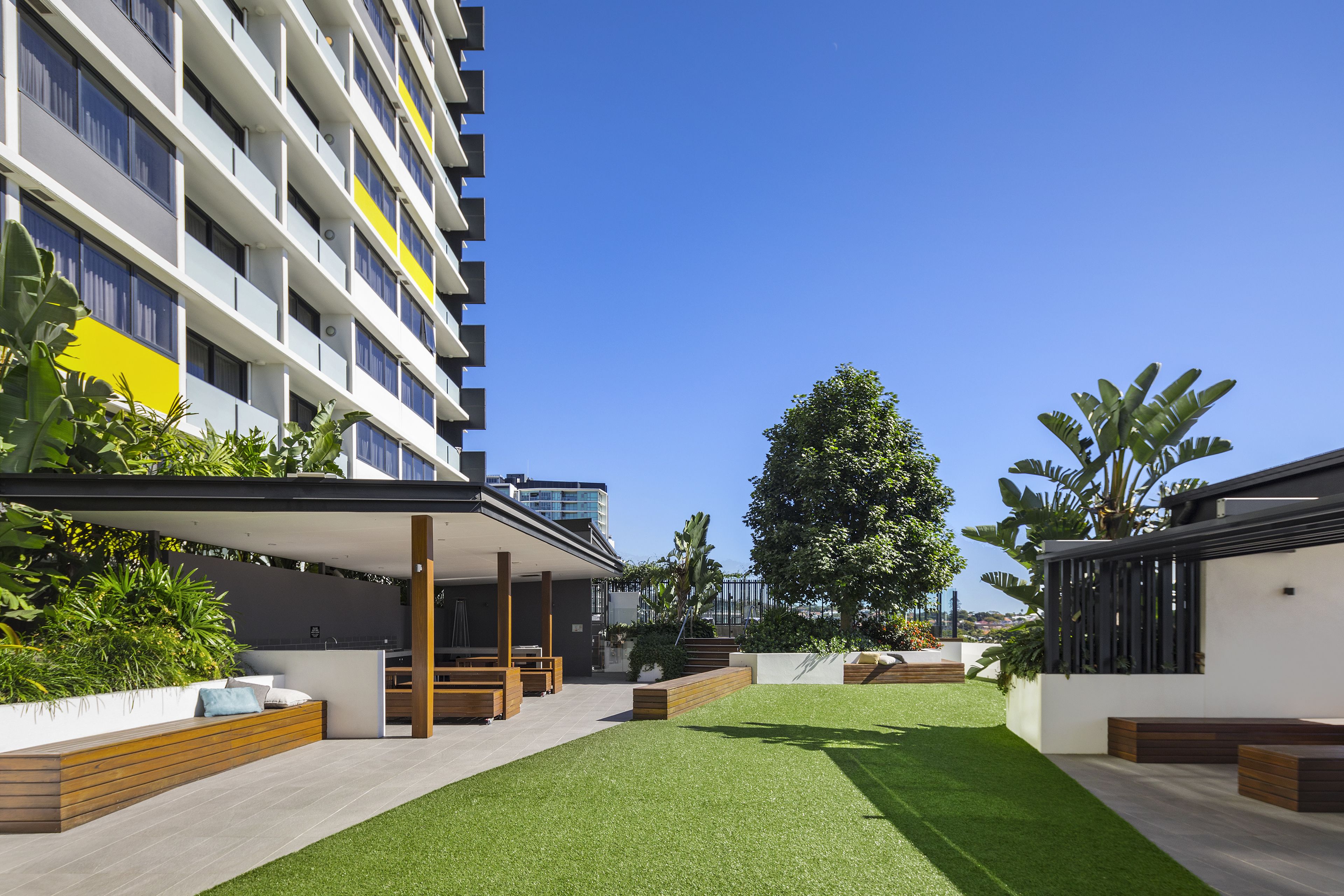 A modern apartment building with a rooftop garden, outdoor seating area, and a large tree providing shade.
