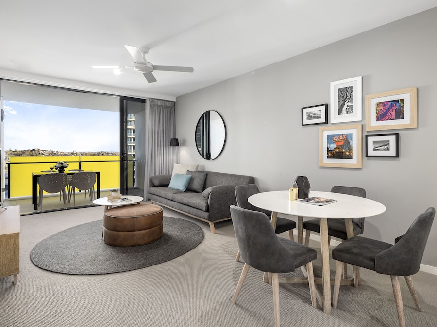 Un salon moderne avec un canapé, une table basse ronde et des fauteuils gris. On y trouve également une table à manger avec des chaises, un téléviseur et un ventilateur de plafond. La pièce est dotée de grandes fenêtres et offre une vue sur l’extérieur.