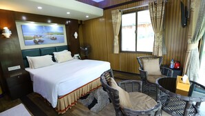 A hotel room with a large bed, a sitting area with wicker chairs, a fan, and a painting on the wall. - Rangauti Resort (Moulvibazar Sadar)