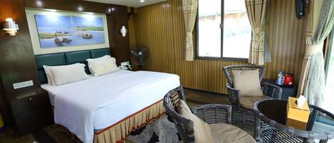A hotel room with a large bed, a sitting area with wicker chairs, a fan, and a painting on the wall.