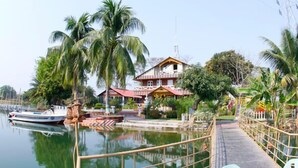A house by a calm water body with a boat docked nearby, surrounded by lush greenery and palm trees. - Rangauti Resort (Moulvibazar Sadar)