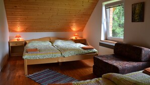 A cozy bedroom with a sloped ceiling, a bed with a green and white striped blanket, two bedside lamps, a window with a view of trees, and a patterned armchair. - Chata Pohoda Slovenský Raj (Spišské Tomášovce)