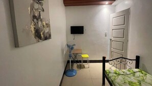 Une chambre à coucher avec un lit, un bureau, une chaise, un ventilateur sur pied et un téléviseur fixé au mur.