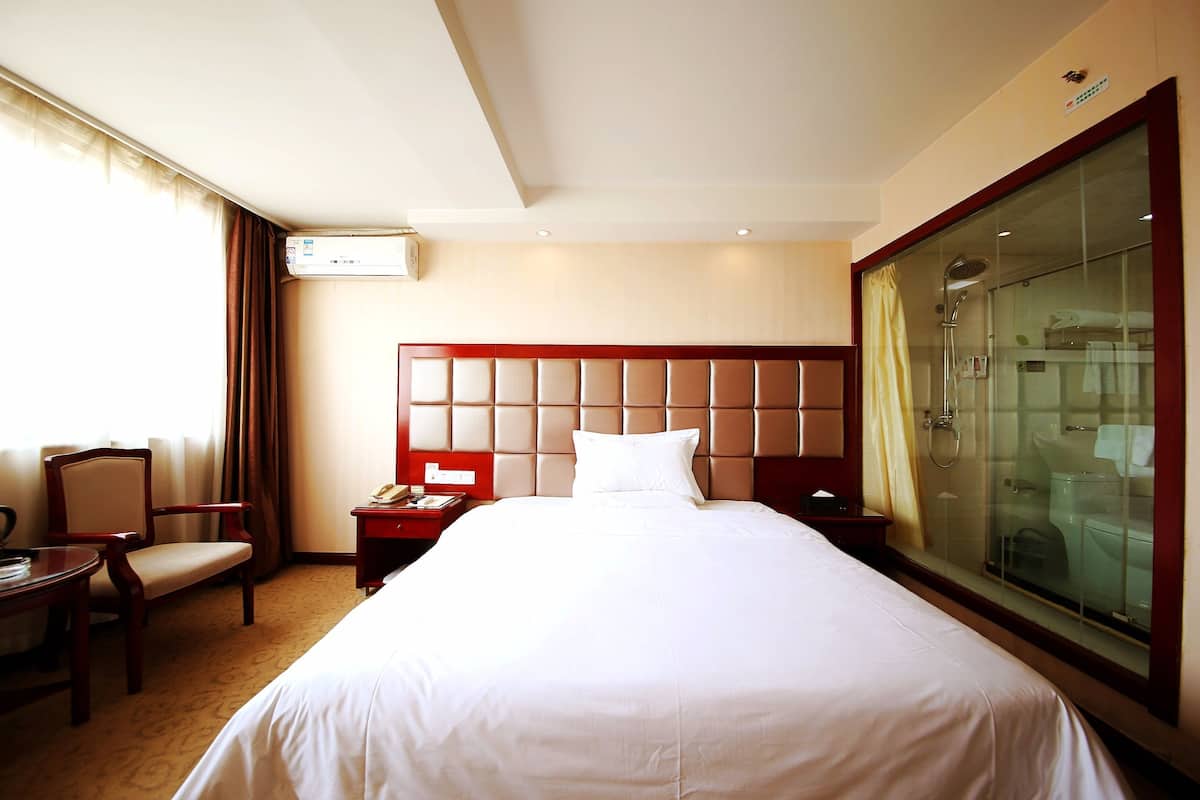 A hotel room with a large bed, a desk with a chair, a bedside table, a telephone, and a glass-enclosed shower area.