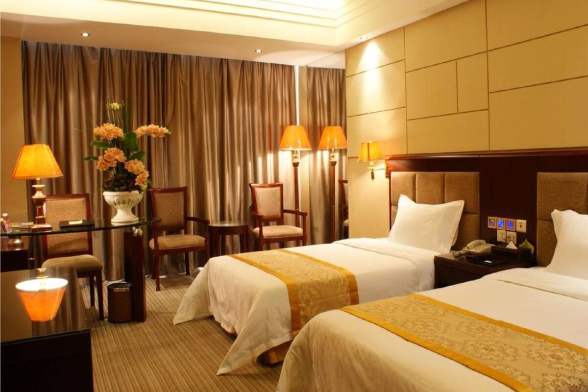 A hotel room with two beds, a desk, chairs, a lamp, and a vase of flowers.