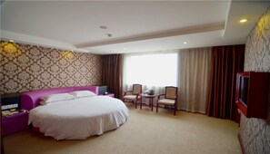 A hotel room with a large bed, a desk with chairs, a television, and a window with curtains.