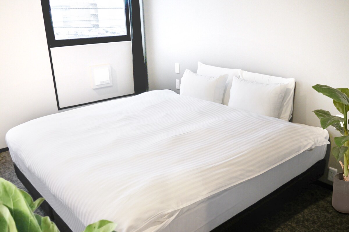 A neatly made bed in a hotel room with a large window, a potted plant, and a wall-mounted light switch.
